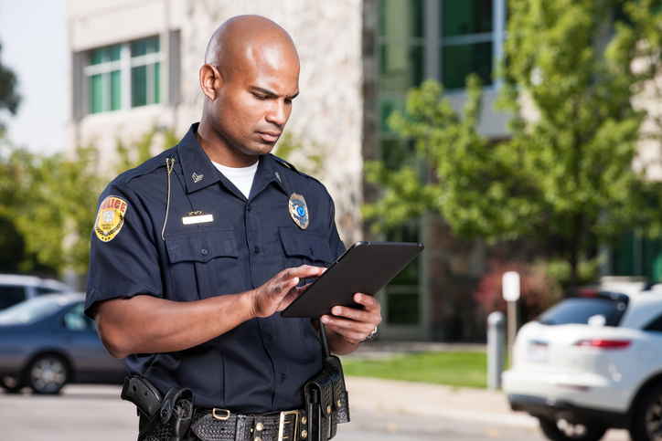 A mid-adult male african american law enforcement officer uses a modern electronic touch screen tablet to enter a citiation or look up information while outdoors.
