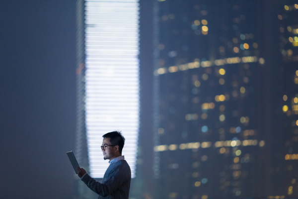 Smart businessman using and looking at digital tablet in city, standing against illuminated financial skyscrapers at night time.